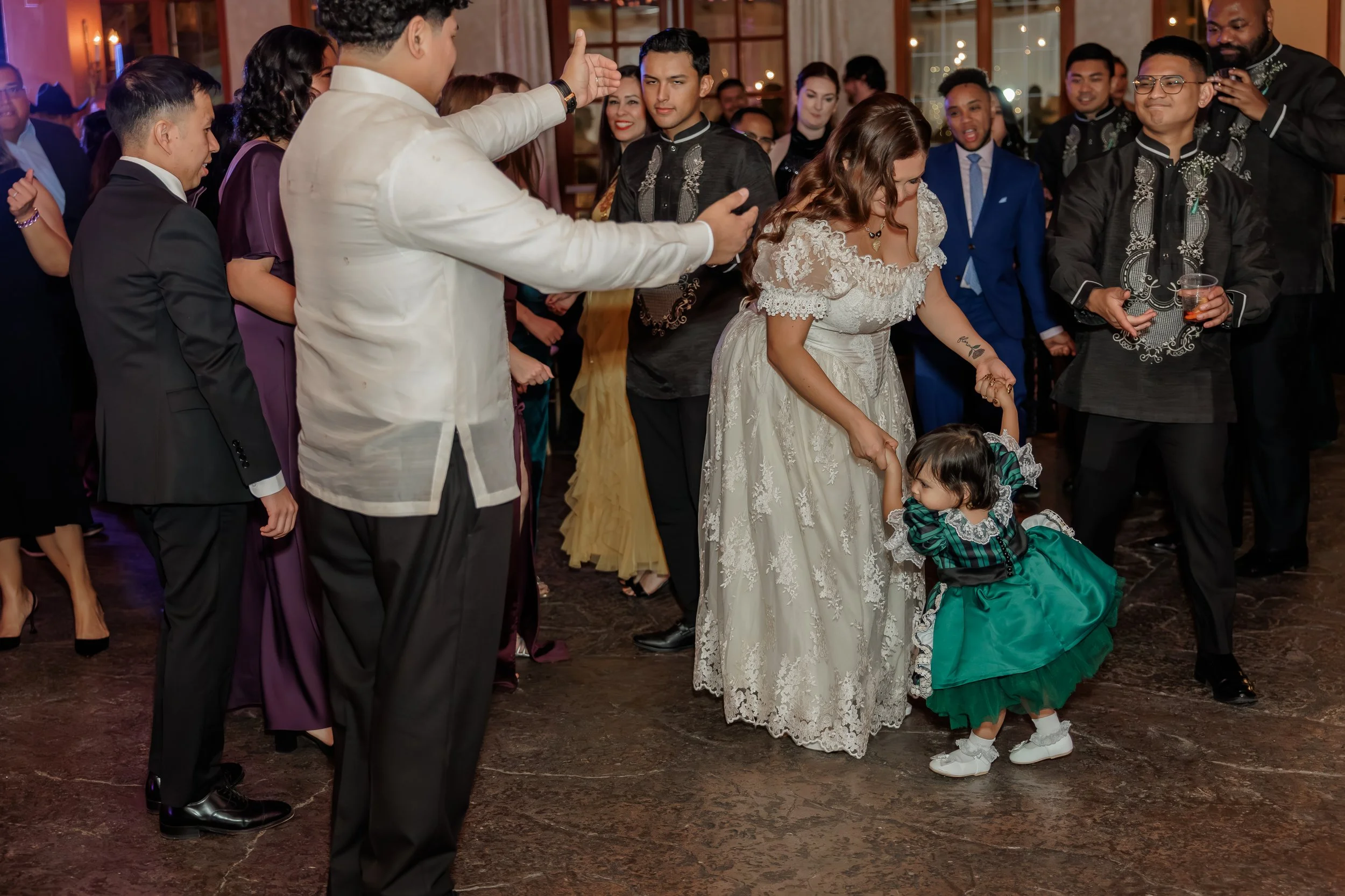 People dancing at a wedding reception, with a bride in a lace dress and two young children dressed in green dancing in front of an audience of well-dressed guests.
