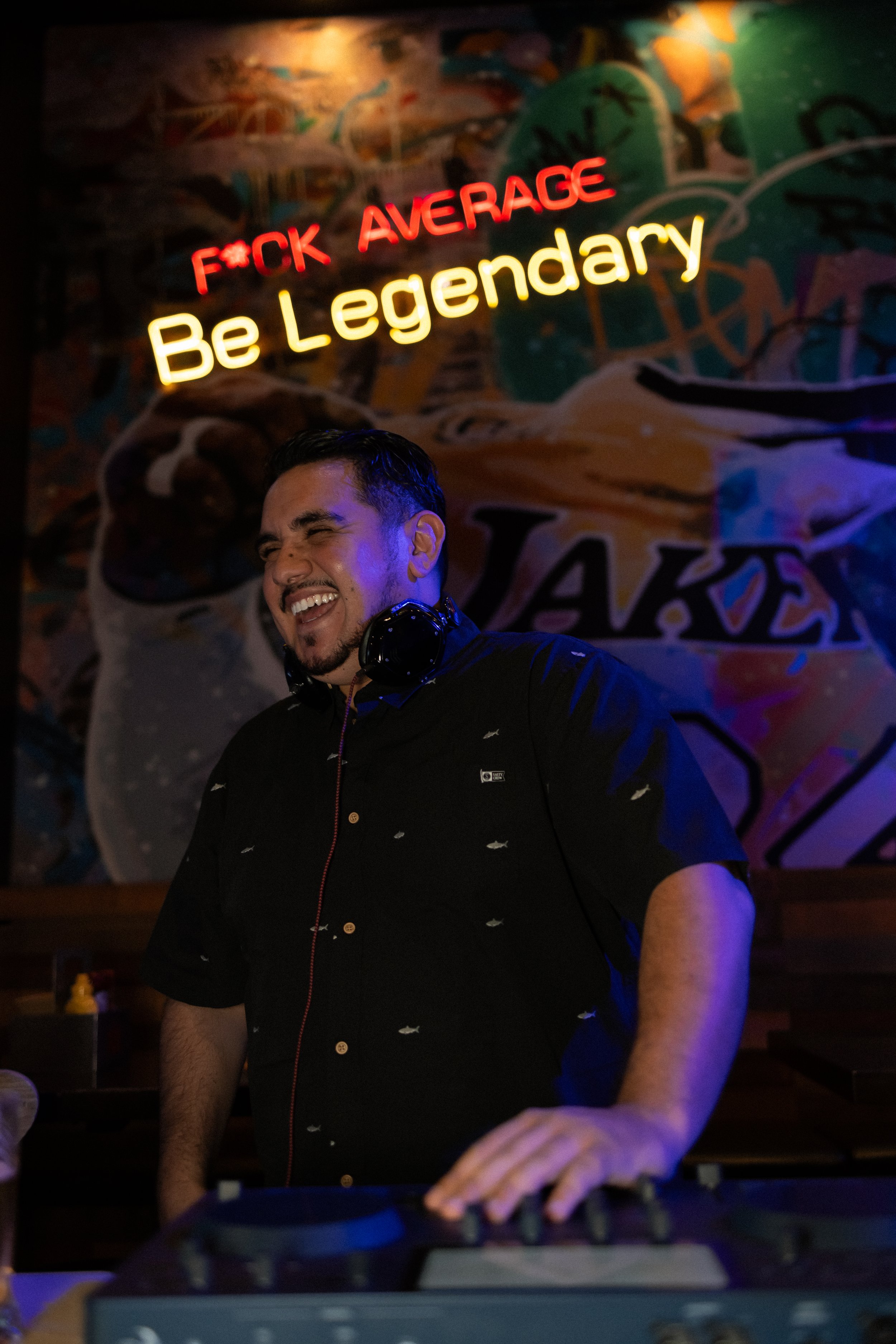 Young man DJ wearing black shirt with fish pattern, smiling and wearing headphones, in front of graffiti wall with neon sign 'F*CK AVERAGE Be Legendary' in red and yellow