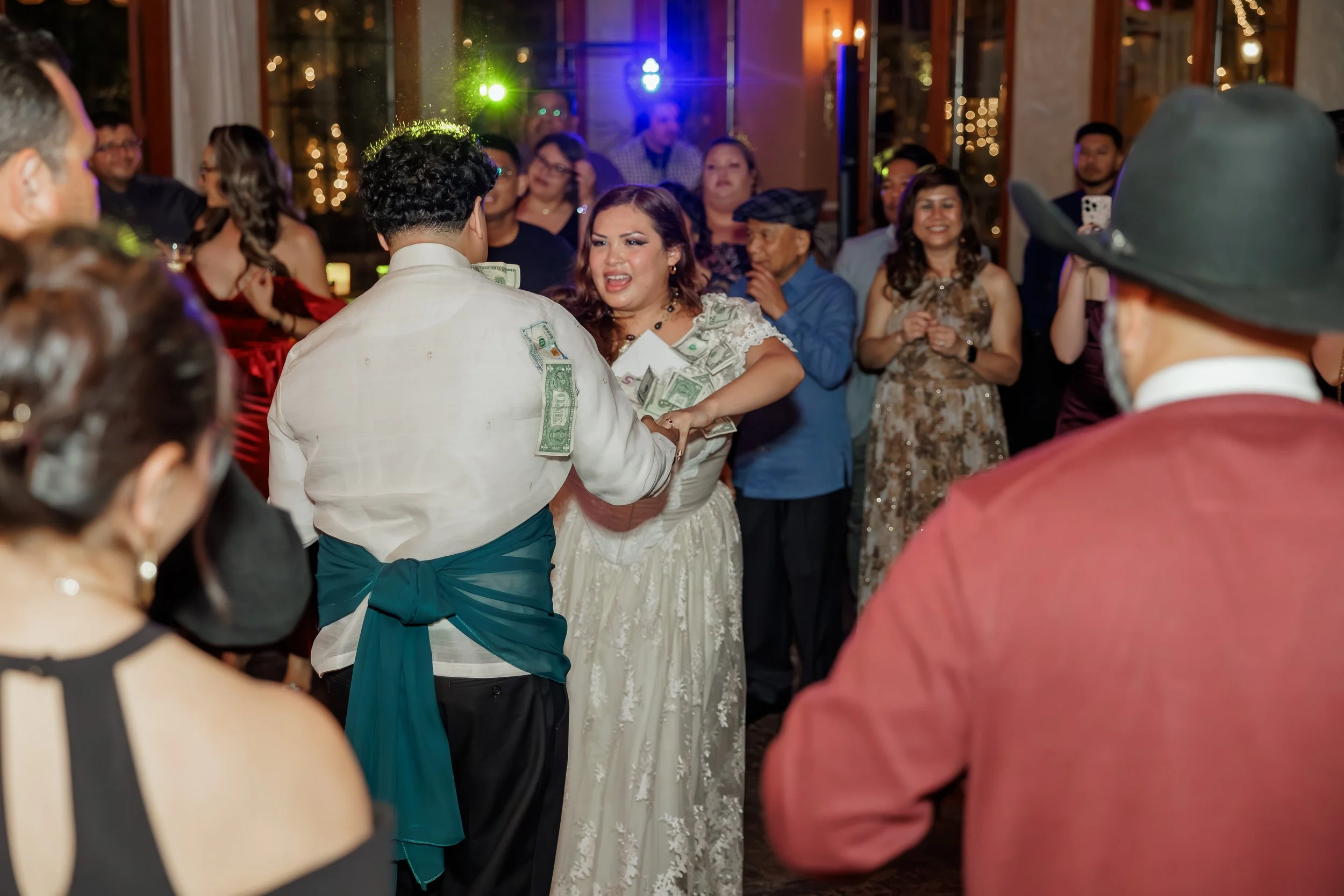 A bride and groom dancing at their wedding reception surrounded by guests, with money pinned to the bride's dress and the groom's suit jacket.
