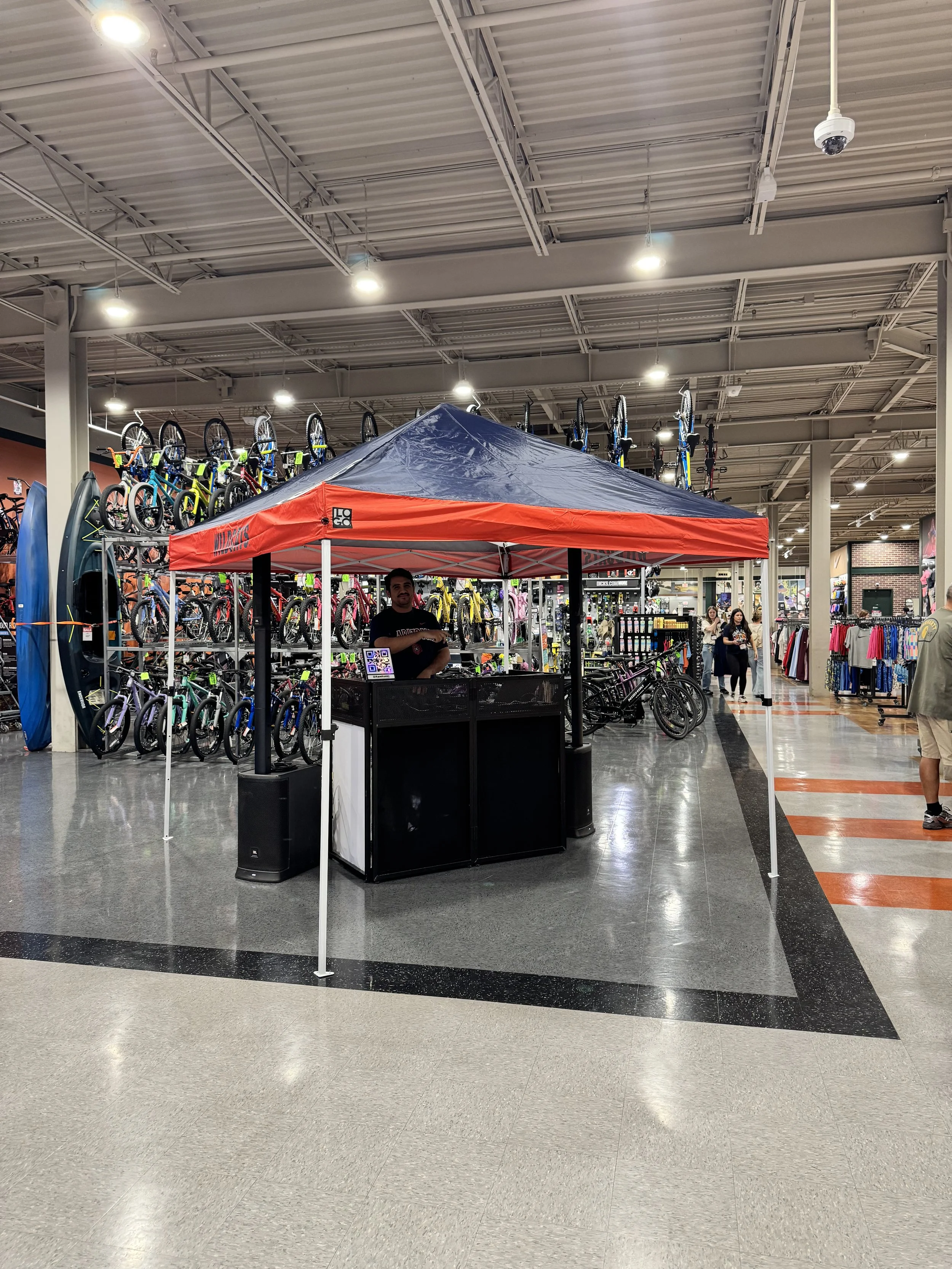 Indoor retail store with a bicycle display area and a small kiosk with a red canopy. The store has bicycles on display on racks and the ceiling has multiple fluorescent lights and security cameras. Customers are shopping in the background.