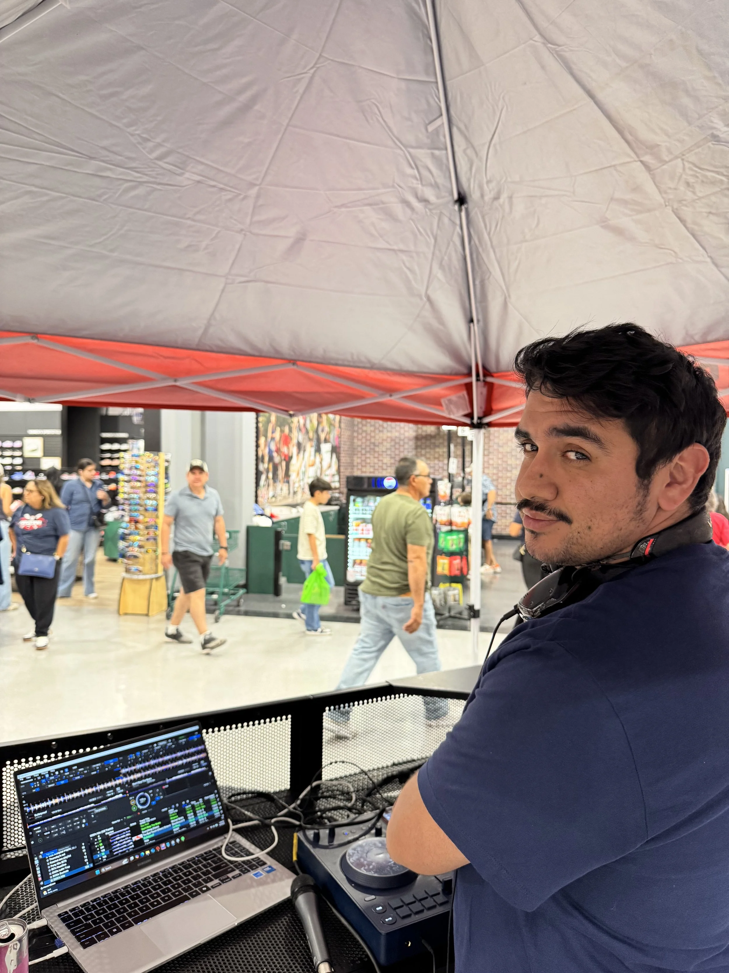 A man with dark hair a is DJing at an event, using a laptop and DJ controller, with people walking in the background under a red canopy at an indoor venue.