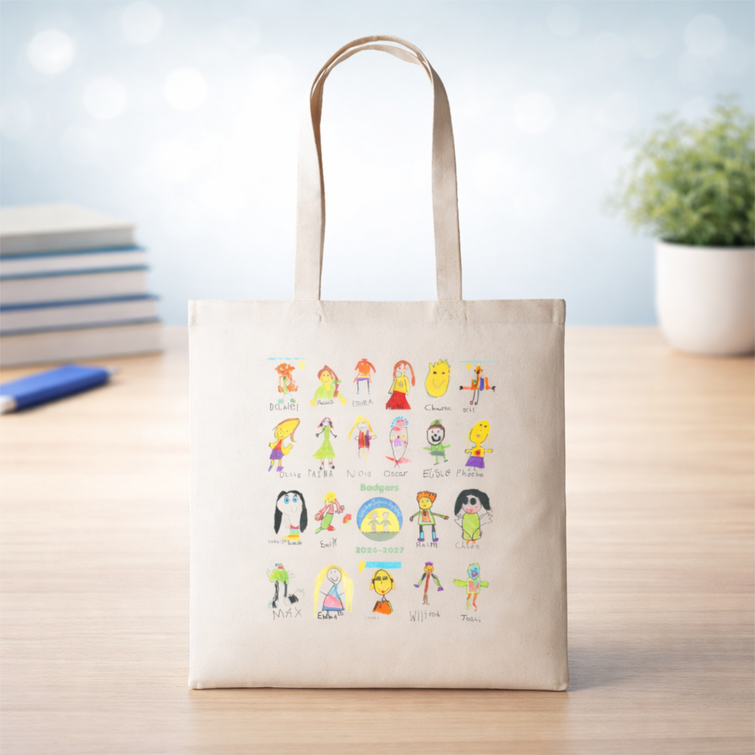 Canvas tote bag with colorful hand-drawn characters and names, including a group labeled "Badgers" and the years 2026-2027, on a wooden table with a blue pen, books, and a potted plant in the background.