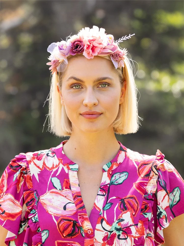 Frau mit Blumenkranz und pinkem Kleid steht im Garten und blickt in die Kamera.
