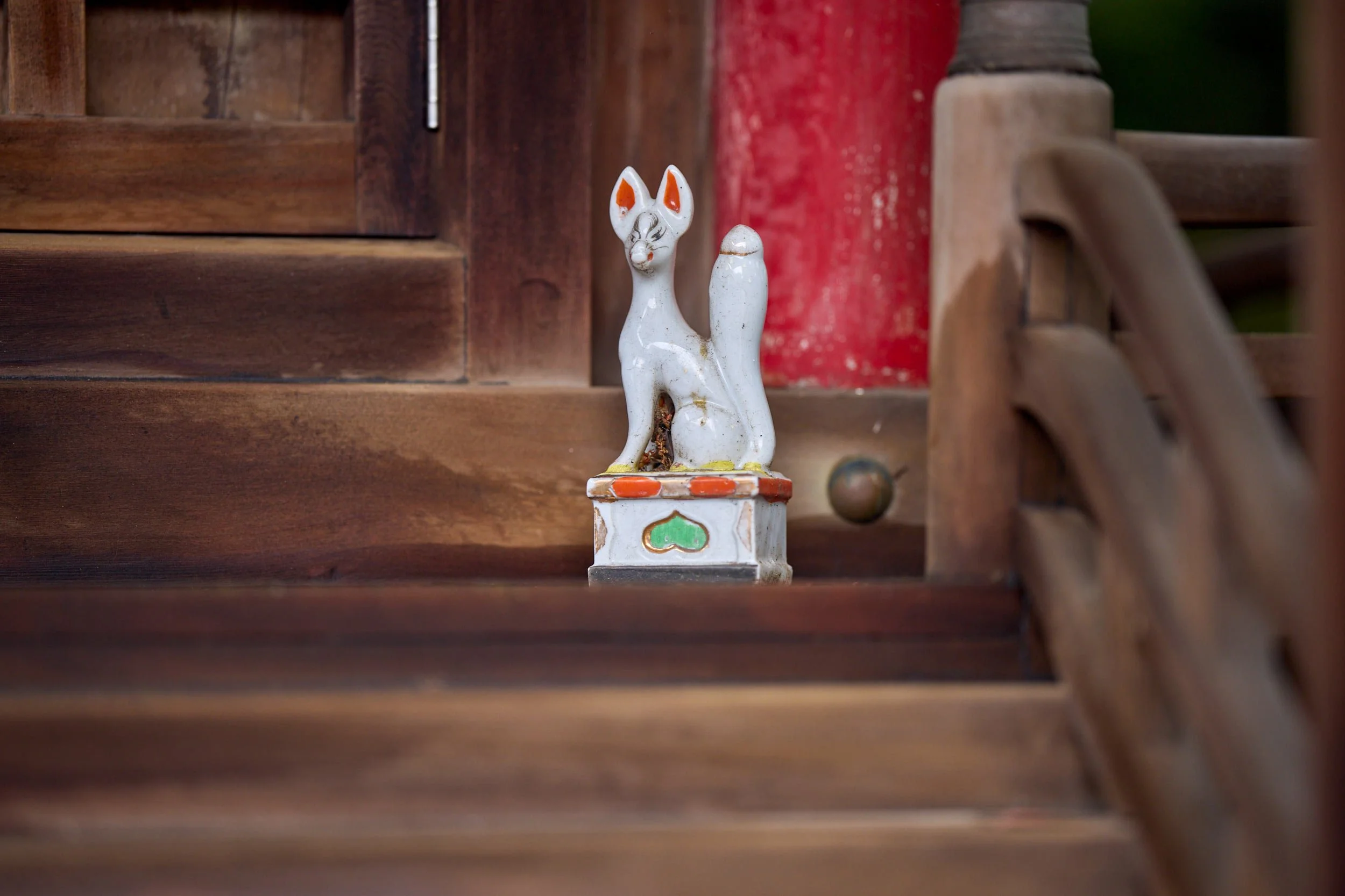 Kleine Kitsune-Fuchsfigur aus Keramik steht auf einem Sockel vor einer Holzstruktur an einem Schrein. Naoshima