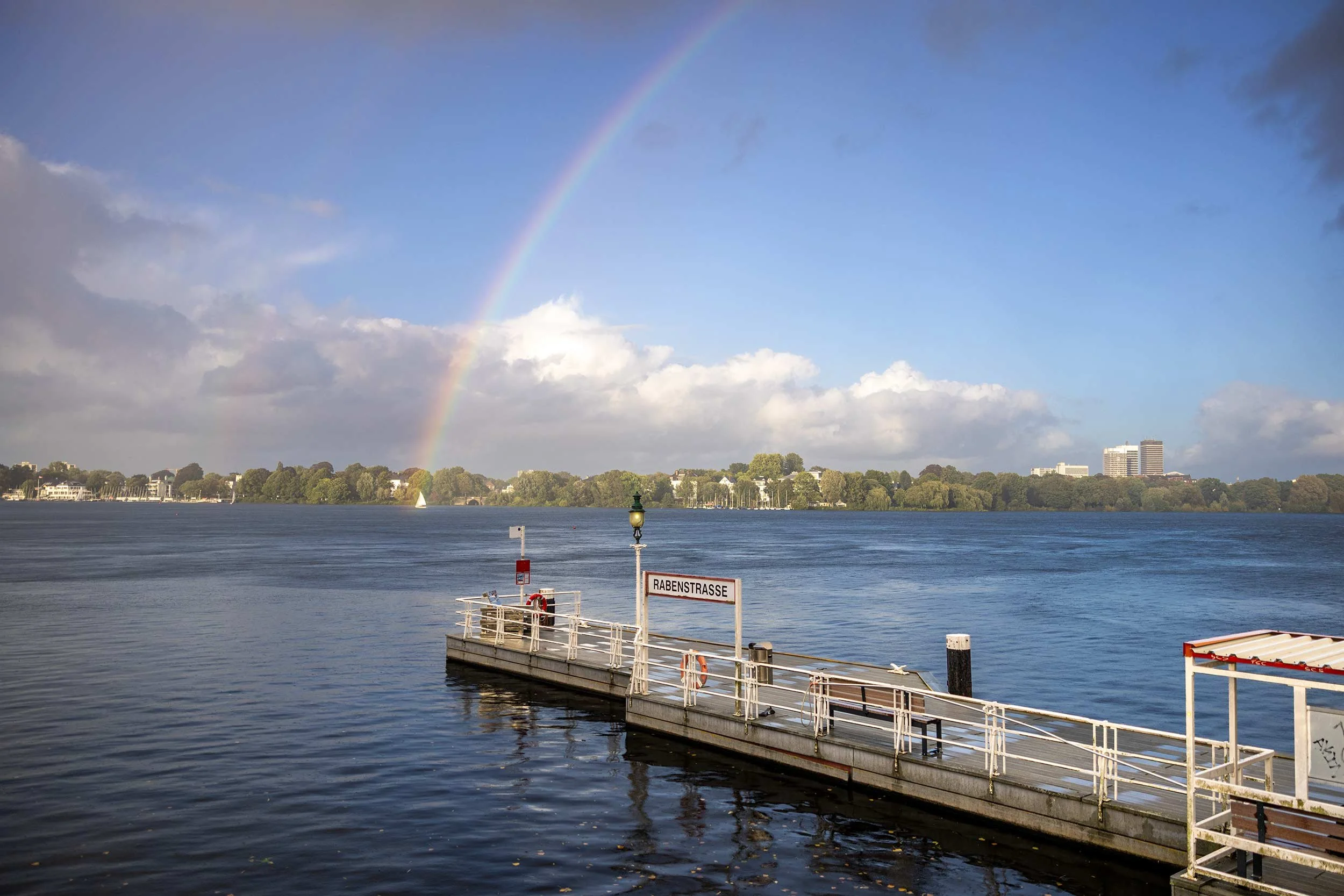 Ein Regenbogen steigt über der Hamburger Außenalster in den aufgehellten Himmel. Im Vordergrund führt der Anleger Rabenstraße diagonal ins Wasser, während sich am gegenüberliegenden Ufer Bäume und Gebäude abzeichnen. Die Aufnahme gehört zur Fotoserie