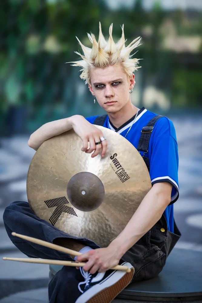 Mitglied der Band III RIØT sitzt mit einem Schlagzeugbecken und Drumsticks vor dem Futurium in Berlin und blickt in die Kamera.