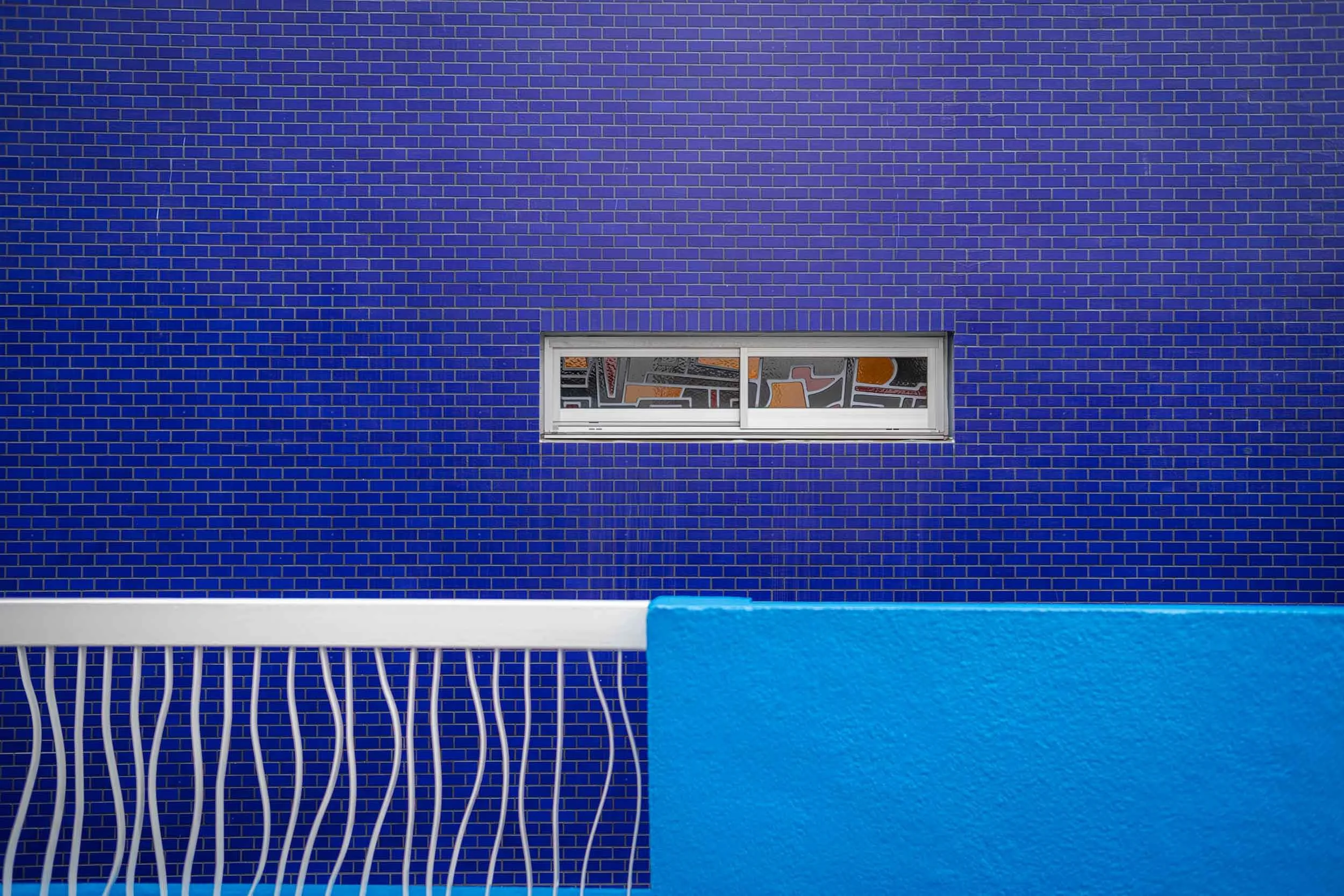 Blaue Fliesenwand mit schmalem Fenster, davor ein weißes Geländer und eine blaue Brüstung.