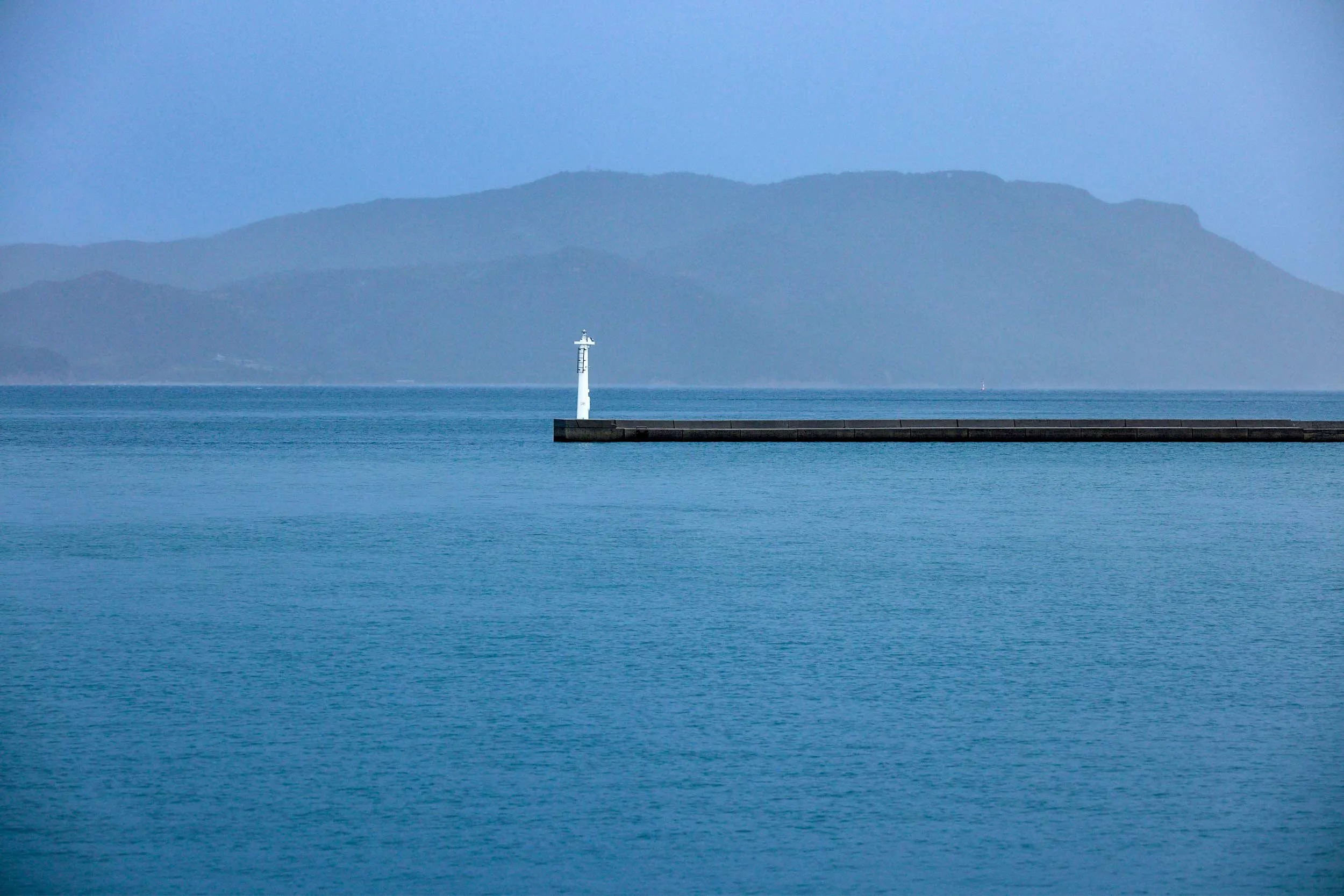 Blick auf das Meer mit langem Wellenbrecher und einem weißen Leuchtturm vor einer Bergsilhouette.