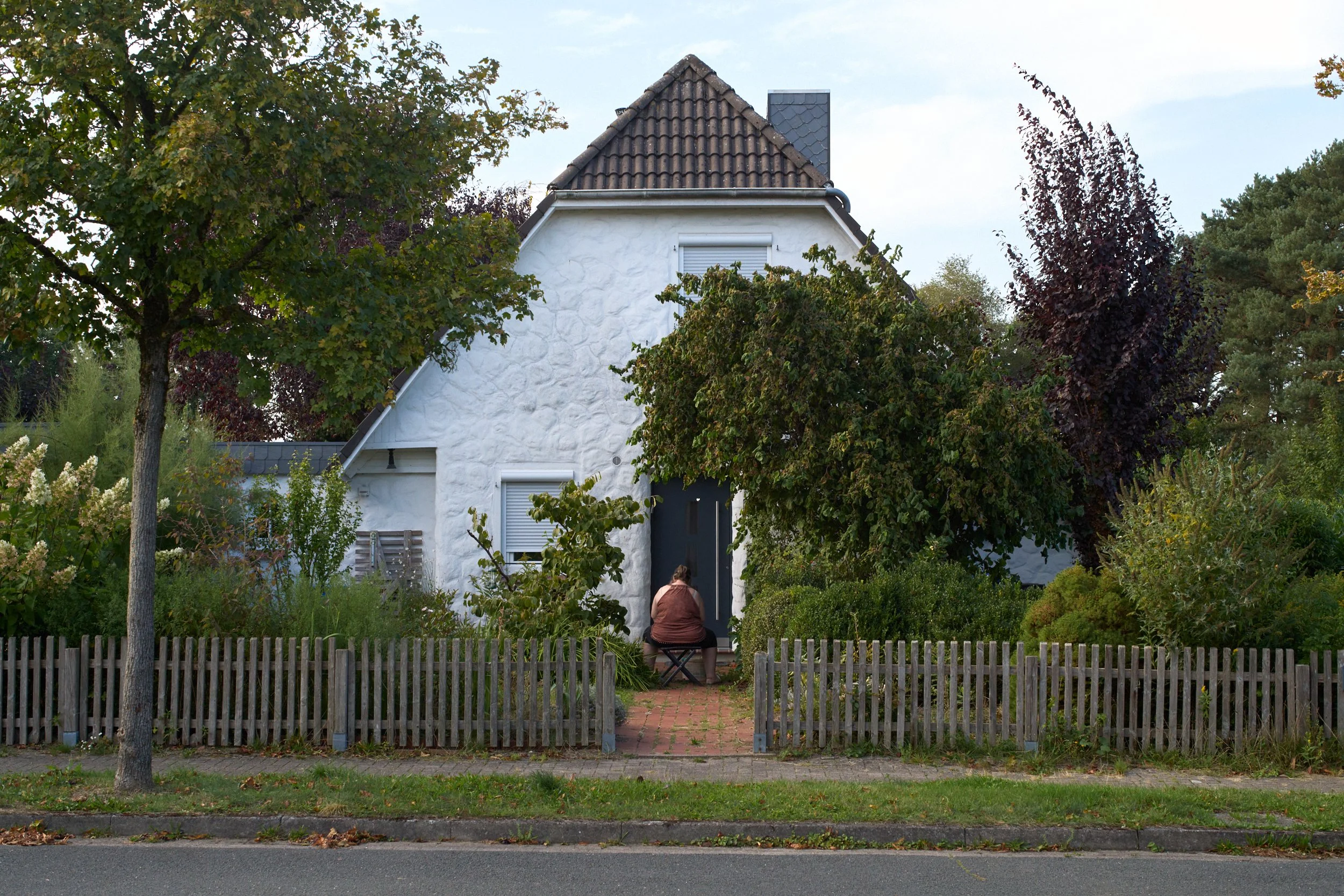 Eine Person sitzt vor dem Eingang eines Wohnhauses in einem bepflanzten Vorgarten.