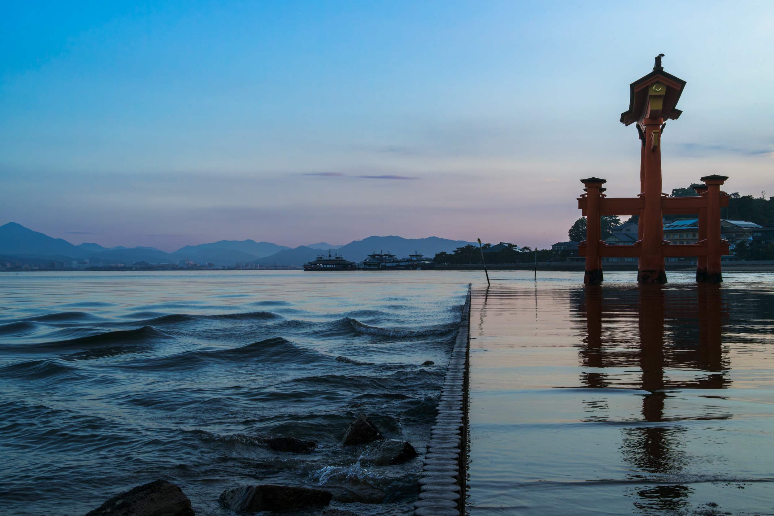 Rotes Torii-Tor steht im flachen Wasser, im Vordergrund Wellen und ein schmaler Damm.