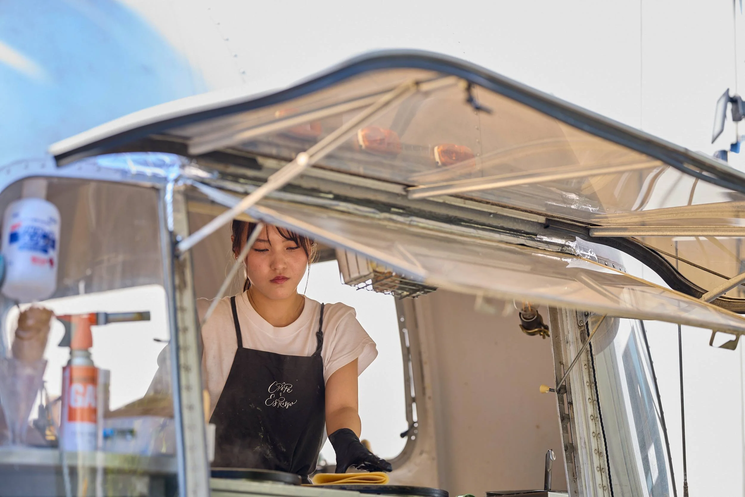 Frau steht in einem Foodtruck in Tokio und bereitet Speisen auf einer Arbeitsfläche zu, sie trägt eine Schürze und Handschuhe, das Dach des Fahrzeugs ist geöffnet.