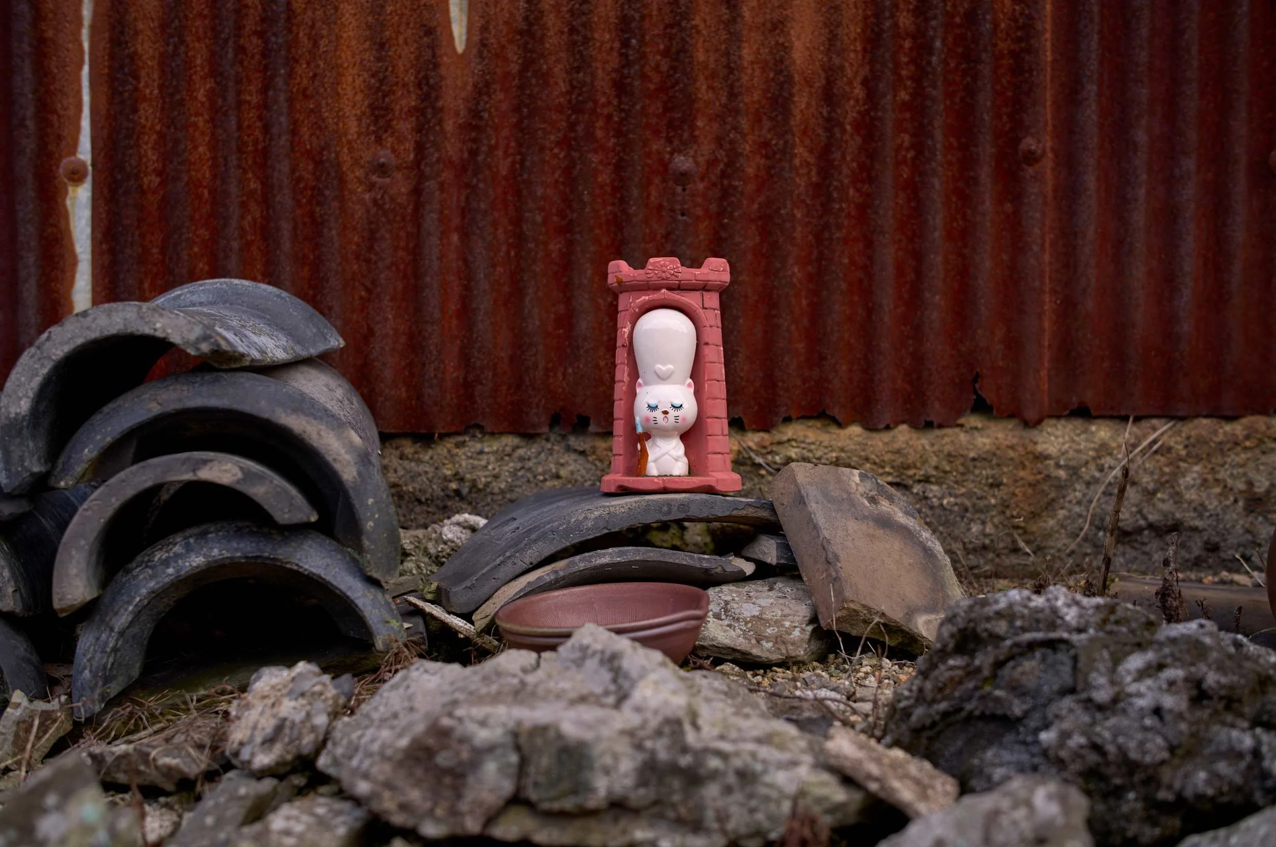 Kleine weiße Figur in einem rosa Rahmen vor einer rostigen Wellblechwand, umgeben von Dachziegeln und Steinen auf Teshima, Japan.