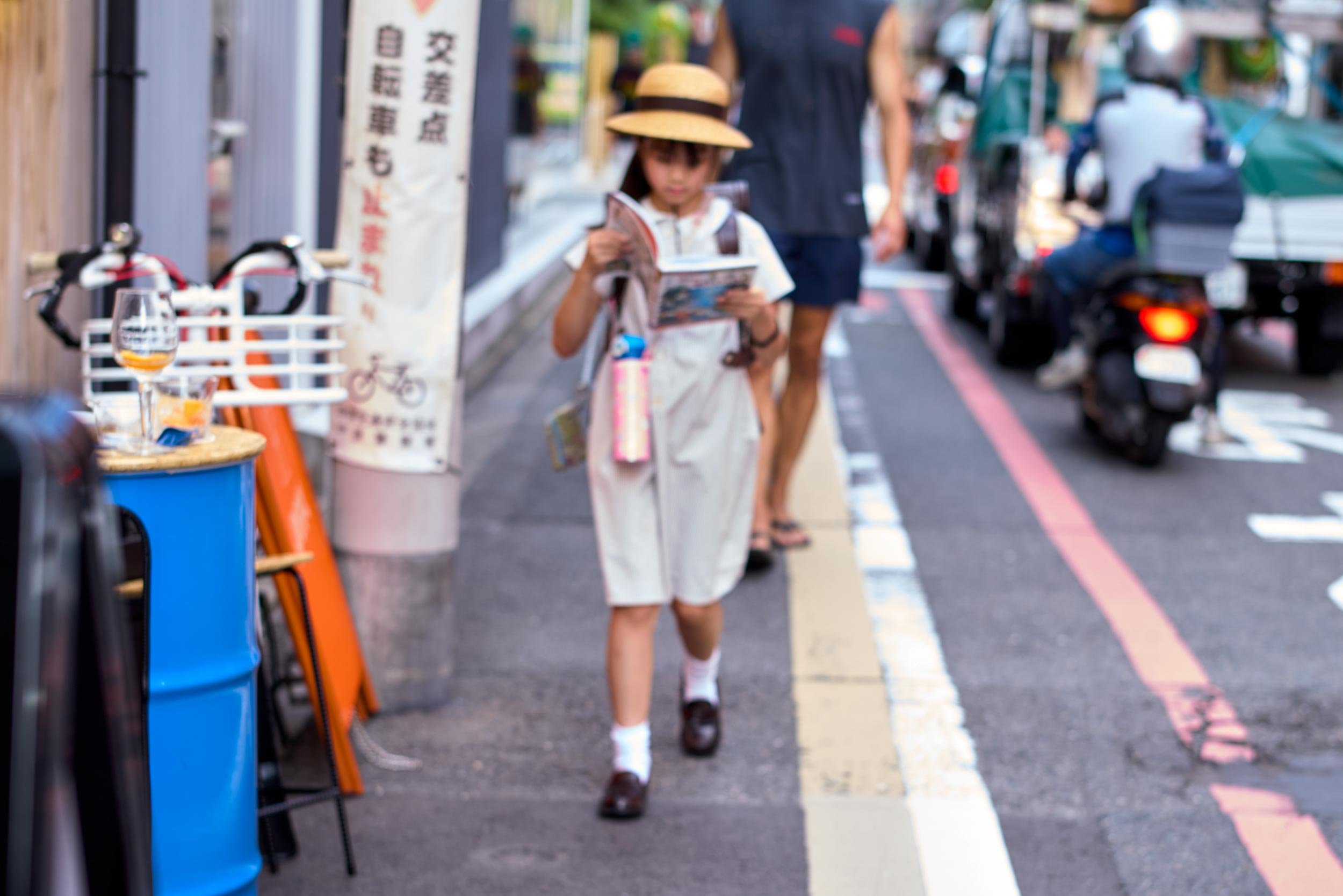 Schulkind mit Strohhut läuft auf einem Gehweg und liest in einem Heft, im Hintergrund Verkehr auf der Straße.