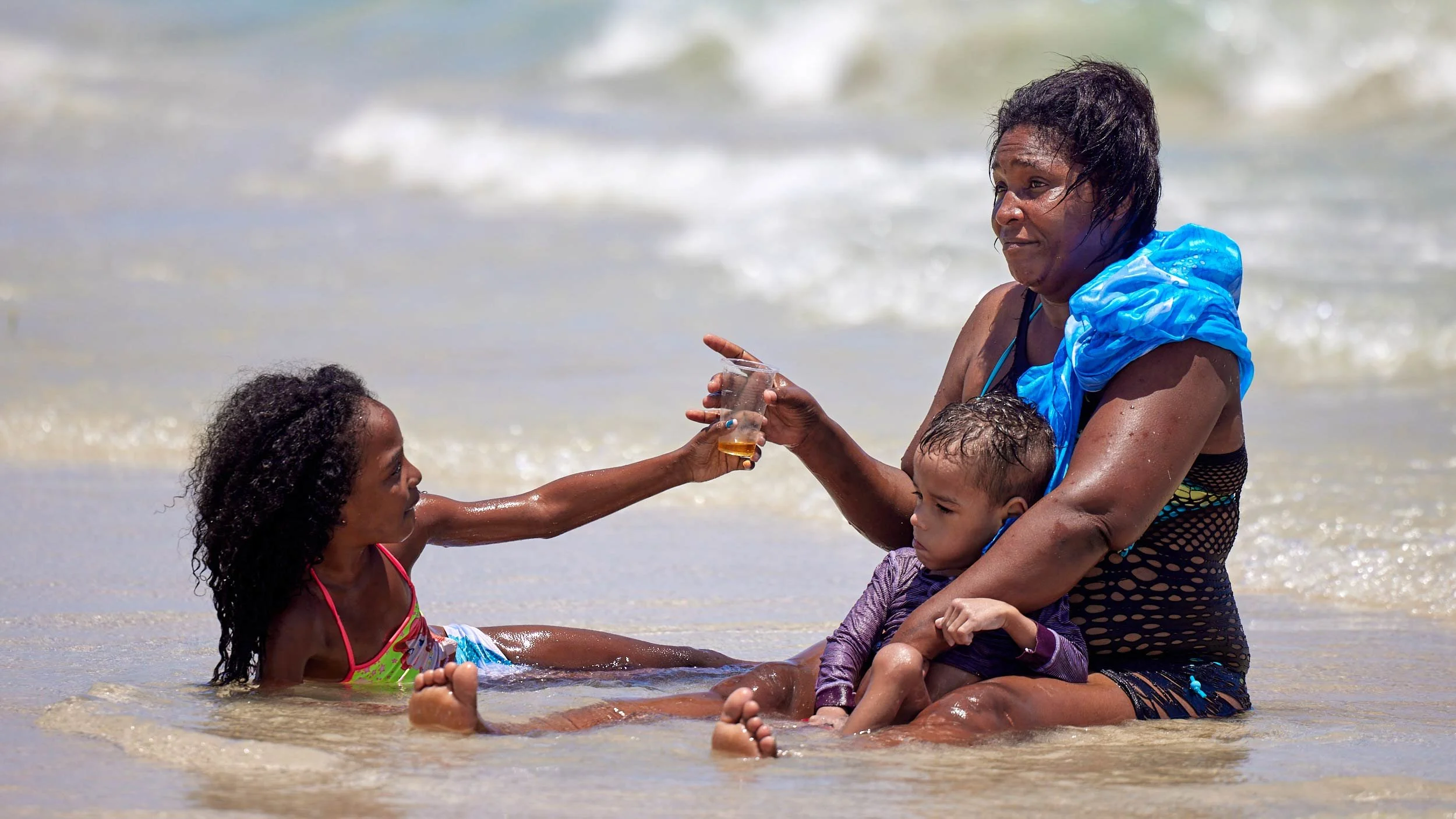 Das Bild zeigt eine intime Szene am Strand, in der eine Frau mit zwei Kindern im seichten Wasser sitzt. Die Mutter hält das jüngere Kind schützend auf ihrem Schoß, während das ältere Mädchen ihr lächelnd einen Cuba anbietet.
