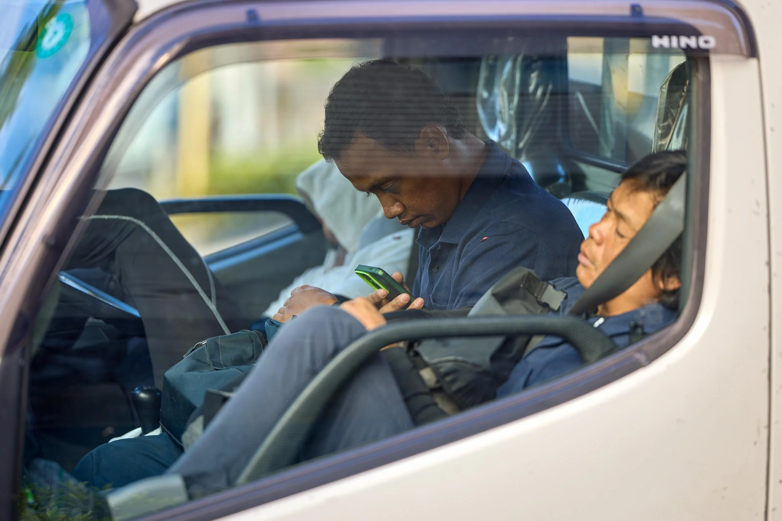 Zwei Männer sitzen in einem Transporter; einer schaut auf ein Smartphone, der andere lehnt mit geschlossenen Augen am Sitz.