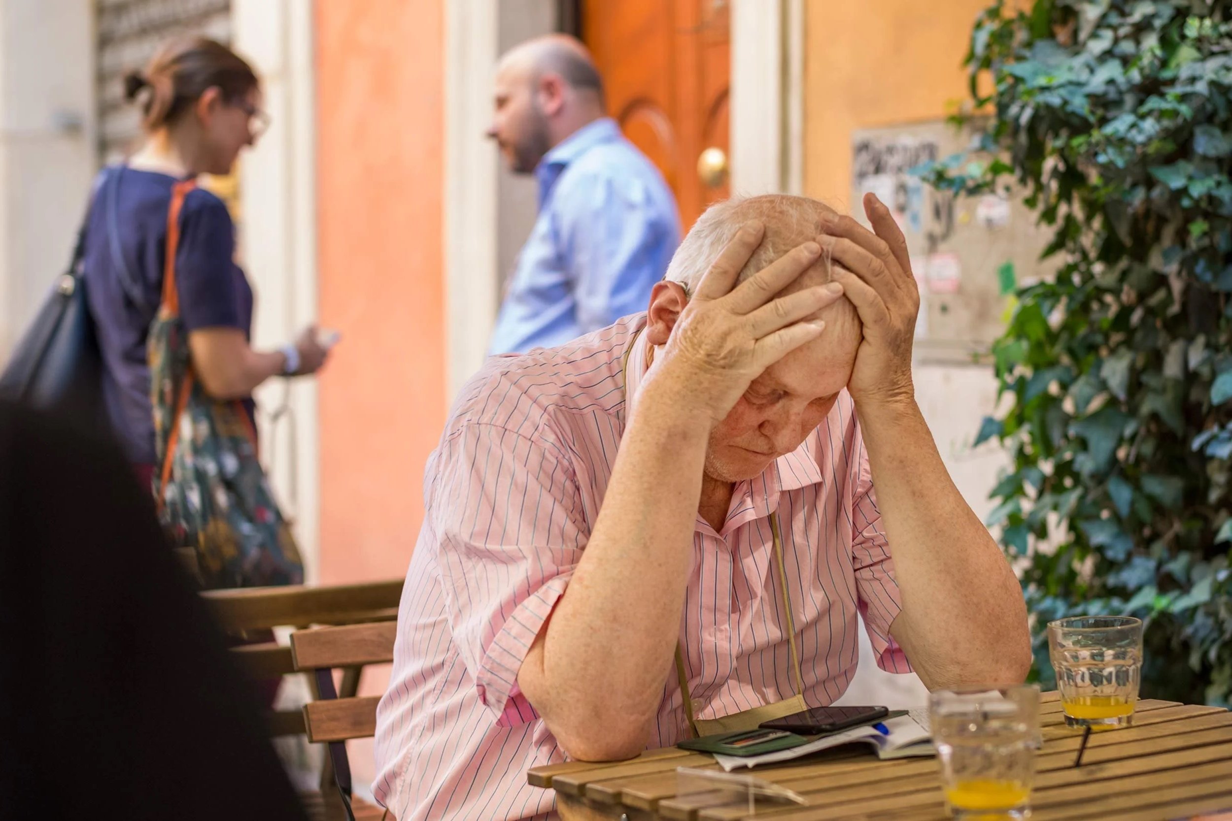 Älterer Mann sitzt an einem Café-Tisch in Rom, stützt den Kopf in die Hände, vor ihm liegen ein Smartphone, ein Notizbuch und zwei Gläser.