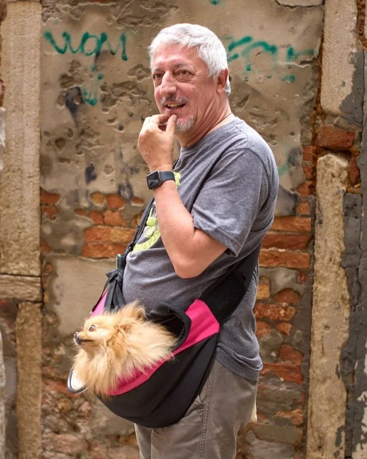 Ein älterer Mann mit grauen Haaren und einem Bart spaziert durch eine enge Gasse in Venedig, Italien. Er trägt ein graues T-Shirt und hat einen kleinen Hund in einer Tasche bei sich. Die Wände der Gasse sind mit Graffiti bedeckt, was eine urbane und 