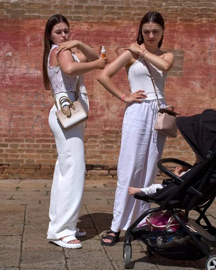 Zwei junge Frauen tragen Sonnenschutzmittel auf ihre Arme auf, während sie in der Sonne stehen. Beide sind in weiße Kleidung gehüllt und stehen vor einer Backsteinwand. Ein Kinderwagen ist im Vordergrund zu sehen, was auf einen Familienausflug hindeu