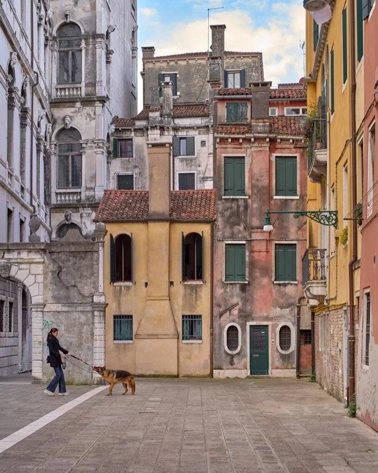 Eine ruhige venezianische Gasse mit hohen, schmalen Häusern in verschiedenen Pastellfarben. Eine Person führt einen Hund an der Leine. Der Platz ist gepflastert und im Hintergrund sind historische Gebäude zu sehen.