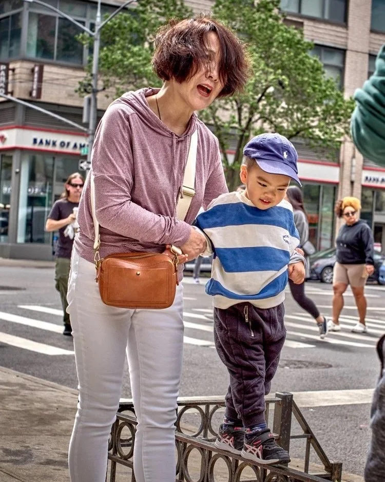 Eine Frau in einer lila Kapuzenjacke hilft einem kleinen Jungen in einem blau-weiß gestreiften Pullover in New York City. Sie stehen auf einem Gehweg vor der Bank of America-Gebäude. Das Bild wurde in Queens aufgenommen.