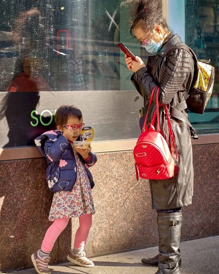 Eine Mutter und ihr kleines Kind stehen vor einem Geschäft in New York City. Die Mutter, die eine Maske und einen schwarzen Ledermantel trägt, schaut auf ihr Smartphone. Sie trägt eine rote Tasche und hat einen Rucksack. Das Kind in bunter Kleidung u