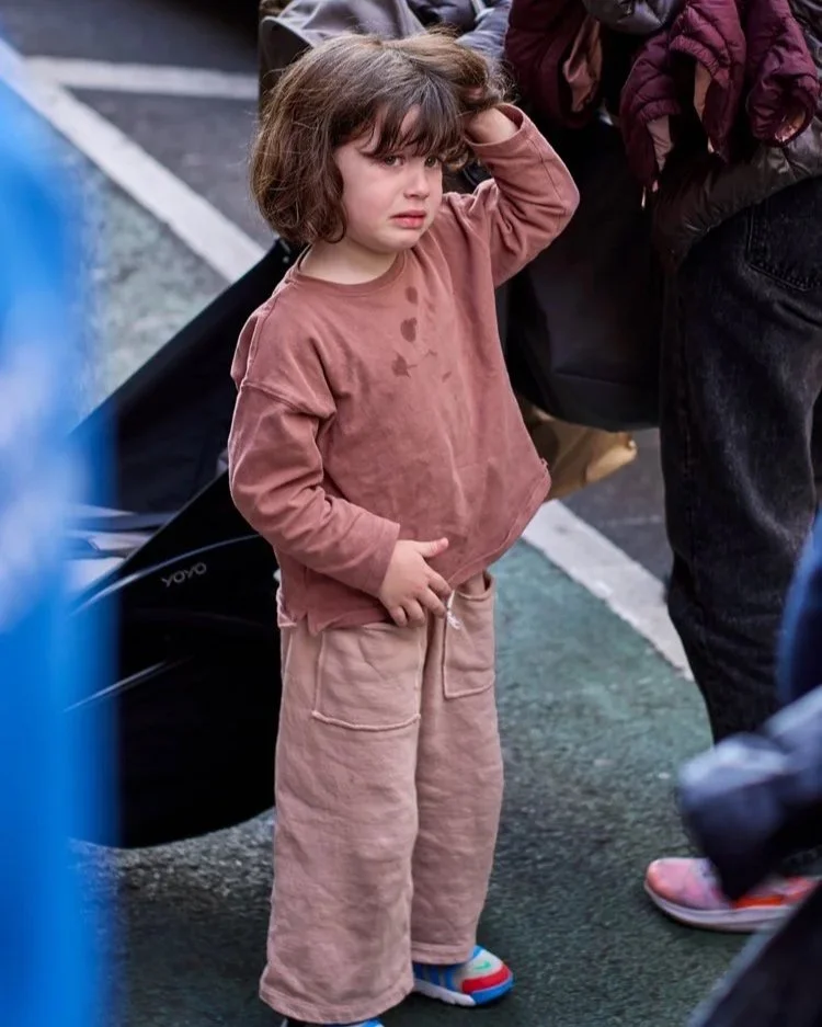 Ein kleines Mädchen in brauner Kleidung steht auf einer belebten Straße in New York City. Sie reibt sich die Augen, ihr Gesichtsausdruck zeigt Erschöpfung und Traurigkeit. Um sie herum sind Erwachsene in Jacken zu sehen, die einen Kinderwagen schiebe