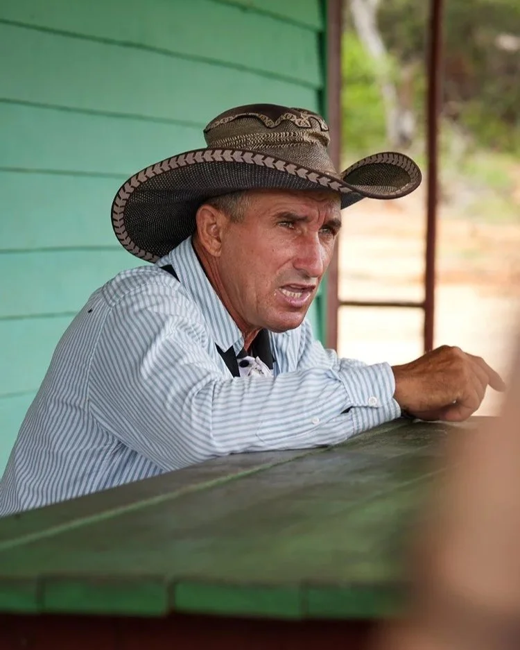 Ein kubanischer Mann mit einem breitkrempigen Hut lehnt an einem hölzernen Tresen, vermutlich vor einem typischen Haus auf dem Land. Sein Gesichtsausdruck ist nachdenklich und erzählt von der harten Arbeit und dem Leben auf dem Land.