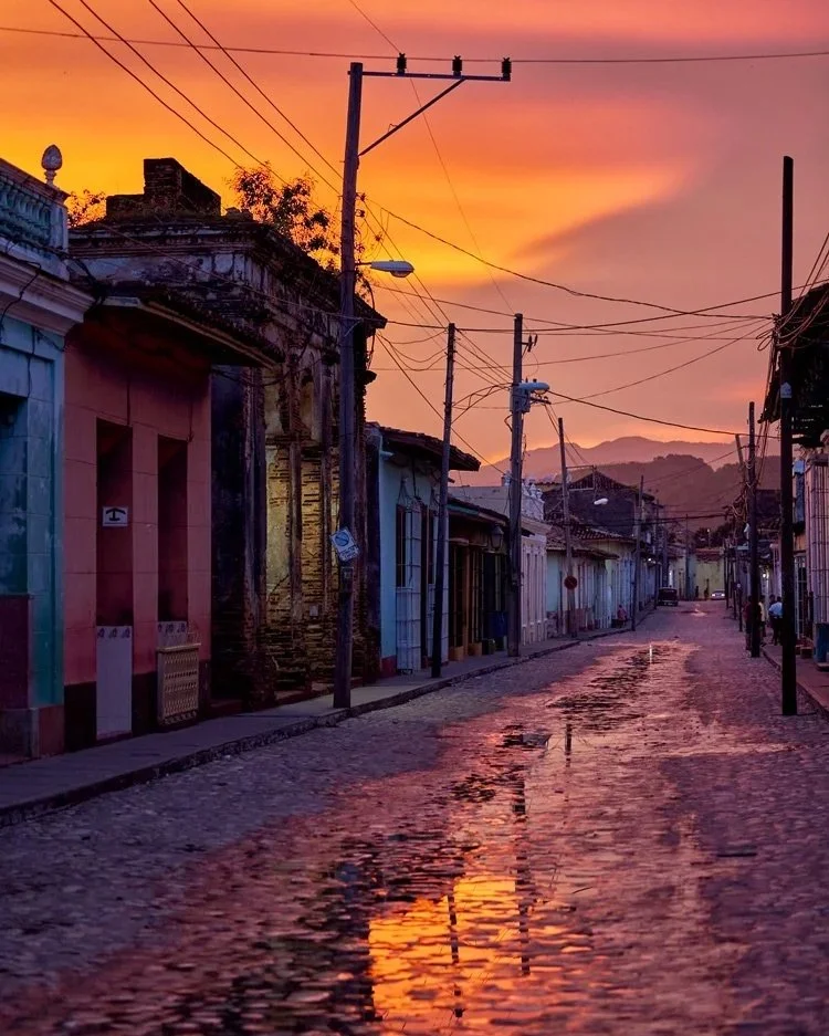Eine malerische Straße in Trinidad, Kuba, bei Sonnenuntergang. Die Straße ist von Wasser bedeckt und spiegelt das intensive, orangefarbene Licht des Himmels wider, was eine warme und nostalgische Atmosphäre schafft.