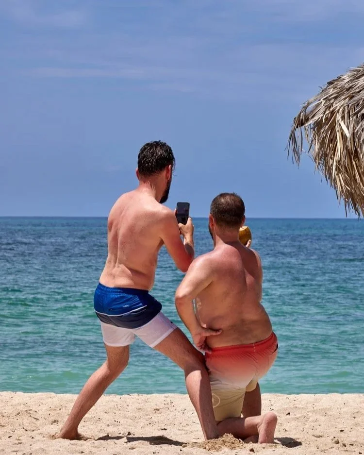Zwei Männer am Strand von Kuba machen Fotos voneinander. Sie stehen im Sand nahe einer Strohhütte und blicken auf das klare blaue Wasser.