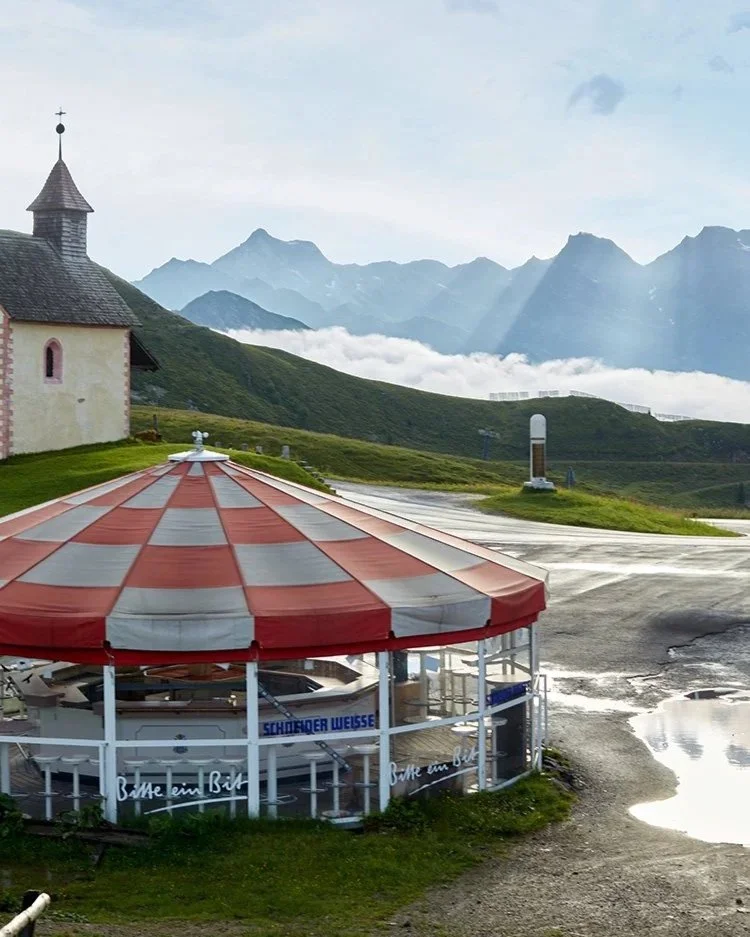 Bildbeschreibung: Rot-weißer Pavillon in den Bergen
Ein rot-weißer Pavillon neben einer kleinen Kirche in einer hochgelegenen Berglandschaft. Die majestätischen Alpen im Hintergrund und Nebelschwaden sorgen für eine malerische Atmosphäre. Standort: G