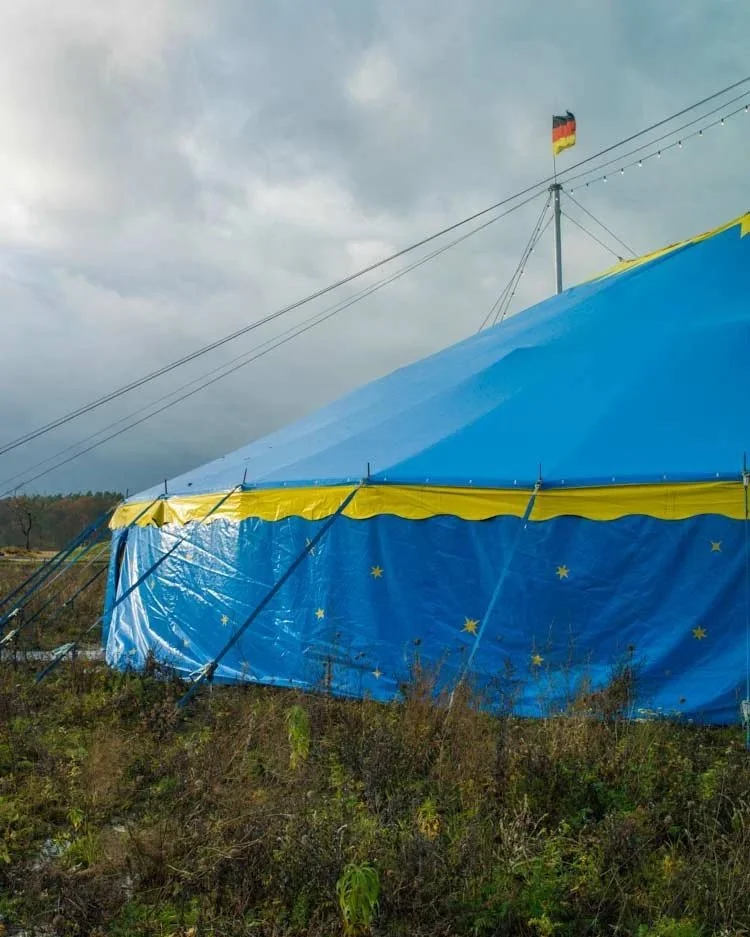 Ein großes blau-gelbes Zirkuszelt mit einer deutschen Flagge auf der Spitze, umgeben von einer natürlichen Landschaft. Die Szene zeigt den Kontrast zwischen dem Zelt und der weiten, grünen Umgebung.