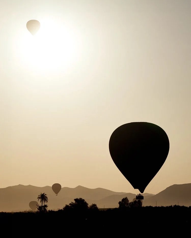 Heißluftballon in der Morgensonne: Vor der goldenen Sonne schwebt ein Heißluftballon über der Wüstenlandschaft Marokkos. Die Silhouetten der Ballons und der Berge erzeugen eine friedliche und stimmungsvolle Szene. Ideal für Abenteuerlustige und Natur
