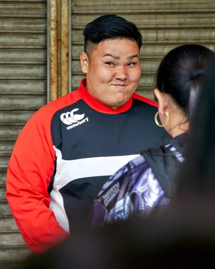 Ein Mann in einem sportlichen Outfit steht vor einem geschlossenen Laden in Tokyo und schaut neugierig in die Kamera. Sein freundliches Lächeln und die lebendige Umgebung spiegeln die gastfreundliche Kultur Japans wider.