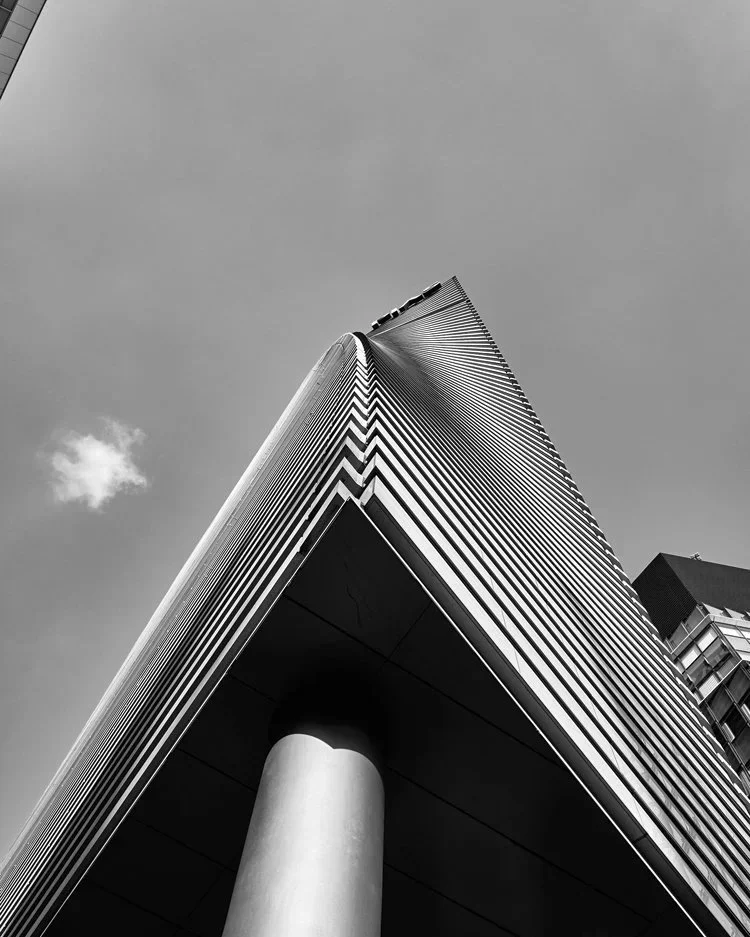 Ein futuristisches Hochhaus mit scharfen Linien und metallischen Details, das in den Himmel ragt. Der Stil ist minimalistisch und erzeugt durch den Schwarz-Weiß-Effekt eine dramatische, monumentale Atmosphäre. Ort: Tokio
