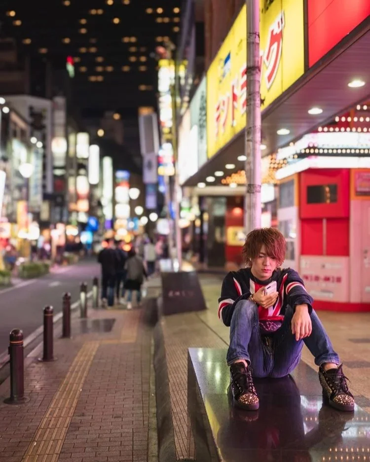  Ein junger Mann sitzt entspannt auf einem Bordstein im belebten Viertel. Um ihn herum leuchten bunte Neonlichter, die typisch für Tokios geschäftiges Nachtleben sind. Die Szene vermittelt eine lässige, städtische Atmosphäre.