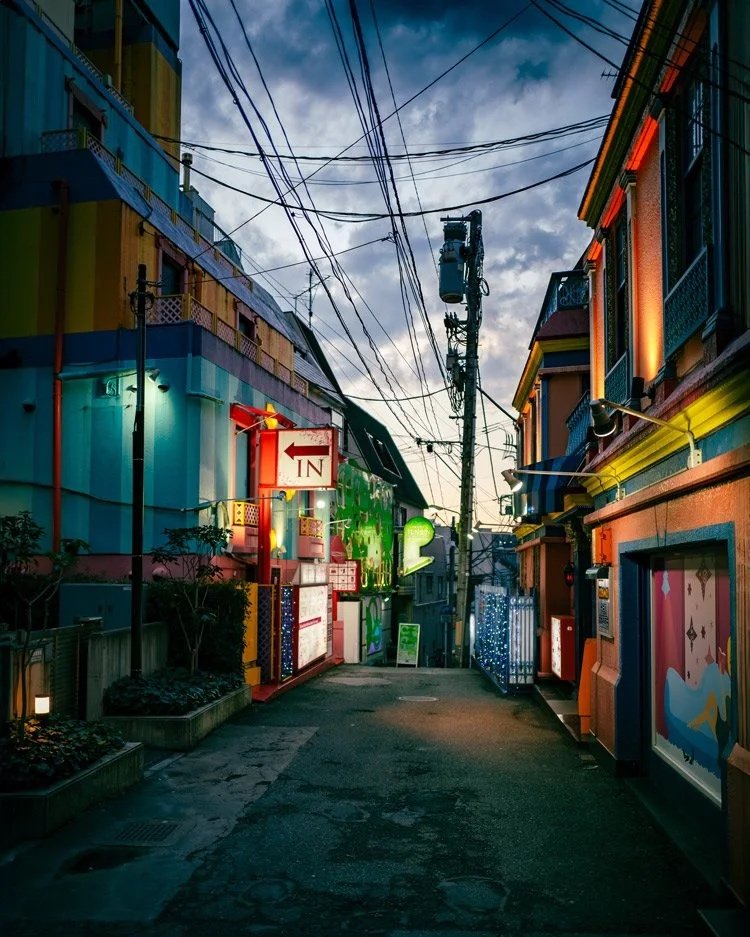 Eine farbenfrohe Gasse in der Dämmerung, beleuchtet von bunten Lichtern und umgeben von überkreuzten Stromleitungen. Die Gebäude sind bunt gestrichen und es herrscht eine ruhige Atmosphäre.
Ortsangabe: Tokyo, Japan.