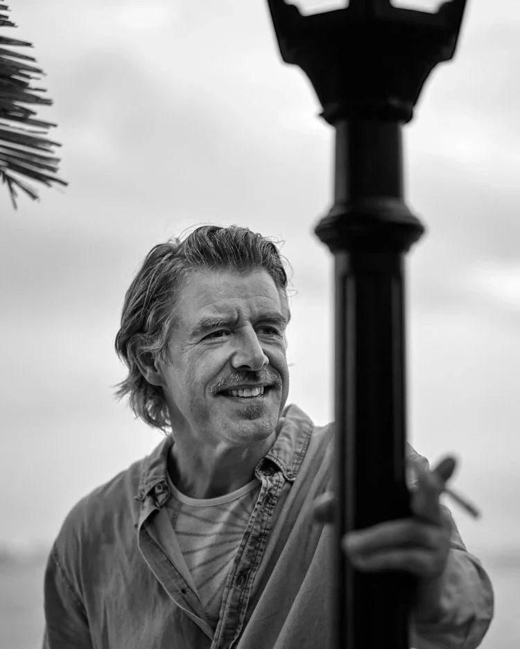 Ein Schwarz-Weiß-Porträt eines lächelnden Mannes im Freien, der eine Zigarette in der Hand hält und sich an einer Laterne anlehnt. Im Hintergrund sind verschwommene Segelboote auf dem Wasser zu sehen.