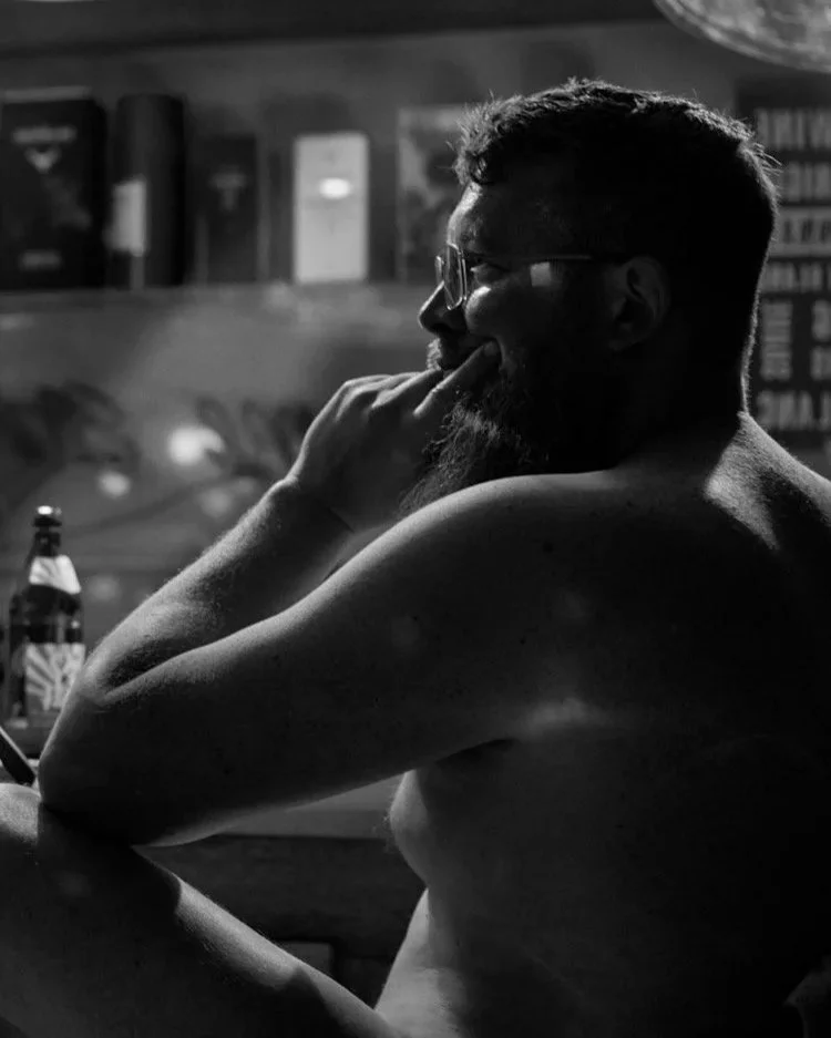 Ein Schwarz-Weiß-Bild eines nackten Mannes, der seitlich zu sehen ist und nachdenklich mit einer Hand an seinem Kinn sitzt. Im Hintergrund sind verschiedene Gegenstände und Regale sichtbar. Das Bild fängt eine ruhige, refle