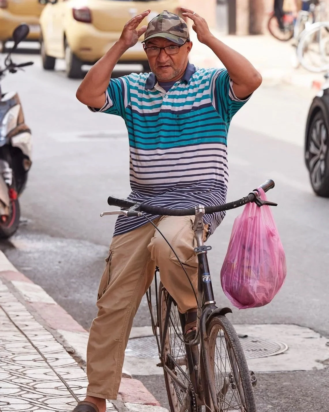 Mann mit Fahrrad: Ein Mann in einem gestreiften Hemd und Sandalen steht neben seinem Fahrrad auf einem Gehweg. In der rechten Hand hält er eine rosa Plastiktüte, während er sich mit der linken Hand die Mütze zurechtrückt. Autos und Motorräder passier