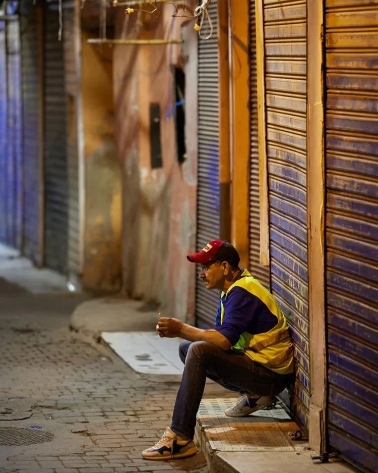Mann sitzt im Basar und raucht: Ein Mann in einer gelben Jacke und roter Kappe sitzt auf dem Bürgersteig des Basars, eine Zigarette in der Hand. Er ist von geschlossenen Metallläden umgeben, die Szene wirkt ruhig und nachdenklich.