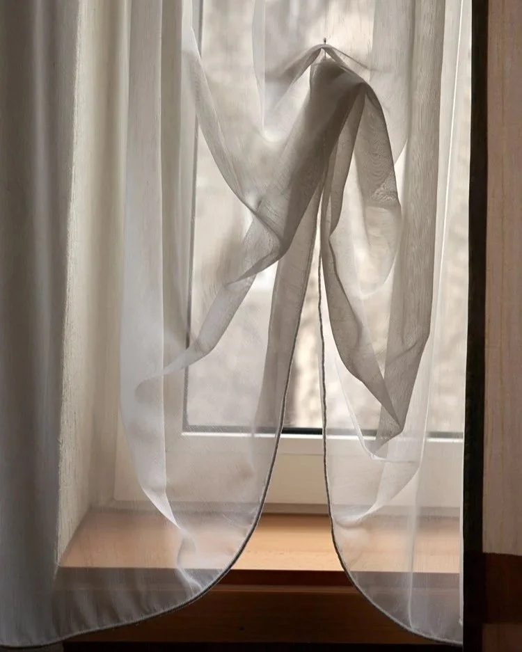 Das Bild zeigt einen zarten, durchscheinenden Vorhang, der leicht geknotet ist und vor einem Fenster hängt, wodurch sanftes Licht in den Raum strömt.