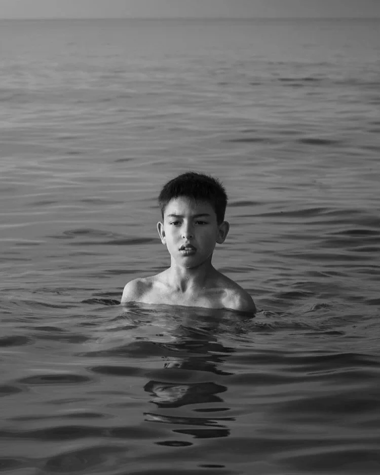 Ein Junge steht bis zu den Schultern im Ozean und schaut ernsthaft in die Kamera. Der Horizont ist im Hintergrund zu sehen, die Stimmung ist ruhig und introspektiv. Ortsangabe: Ostsee