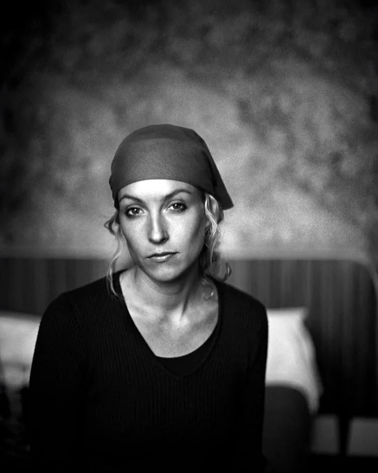 Das Foto zeigt eine schwarz-weiße Aufnahme einer Person mit einem ruhigen Ausdruck. Sie trägt ein Kopftuch und hat lockiges Haar, das leicht herausschaut. Die Person ist von der Brust aufwärts zu sehen, mit einem leicht gesenkten Blick. Im Hintergrun