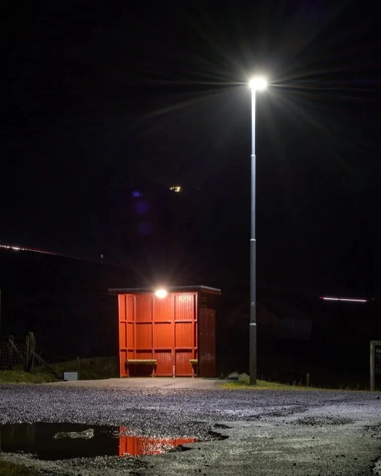 Eine Bushaltestelle auf den Färöer-Inseln bei Nacht. Die Haltestelle ist rot gestrichen und wird von einer Straßenlaterne beleuchtet, die auch den nassen Boden reflektiert. Die Umgebung ist dunkel und ruhig, nur von wenigen Lichtquellen unterbrochen,