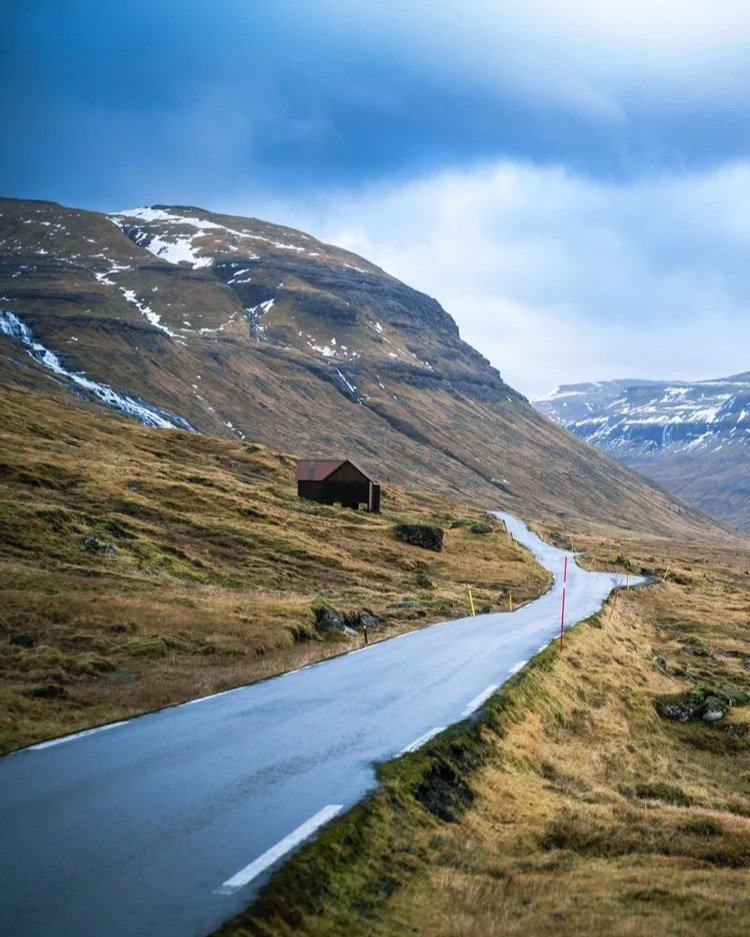 Das Bild zeigt eine einsame Straße, die sich durch die bergige Landschaft Norwegens schlängelt. Im Vordergrund ist eine einfache Hütte zu sehen, während im Hintergrund schneebedeckte Gipfel und sanfte Hügel die wilde, unberührte Natur Norwegens beton