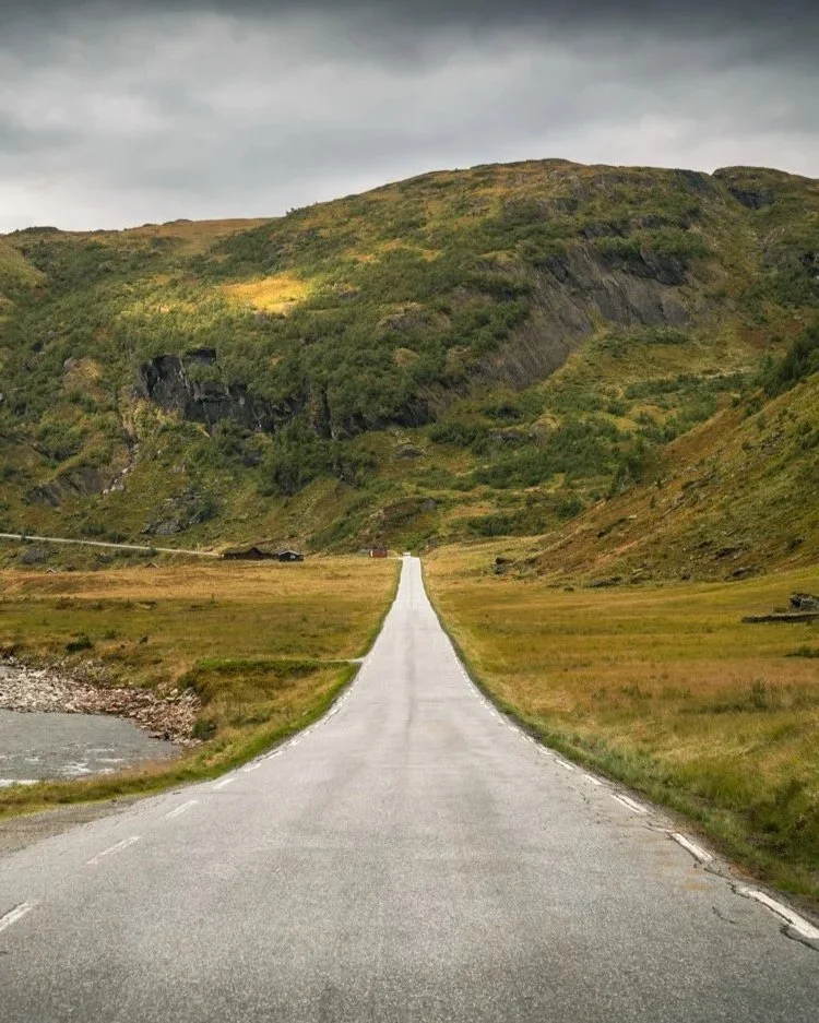 Das Foto zeigt eine lange, gerade Straße, die sich durch eine grüne, hügelige Landschaft in Norwegen erstreckt. Die Straße führt direkt auf einen bewachsenen Hügel zu, über dem sich ein bewölkter, grauer Himmel wölbt. Die Umgebung wirkt ruhig und abg