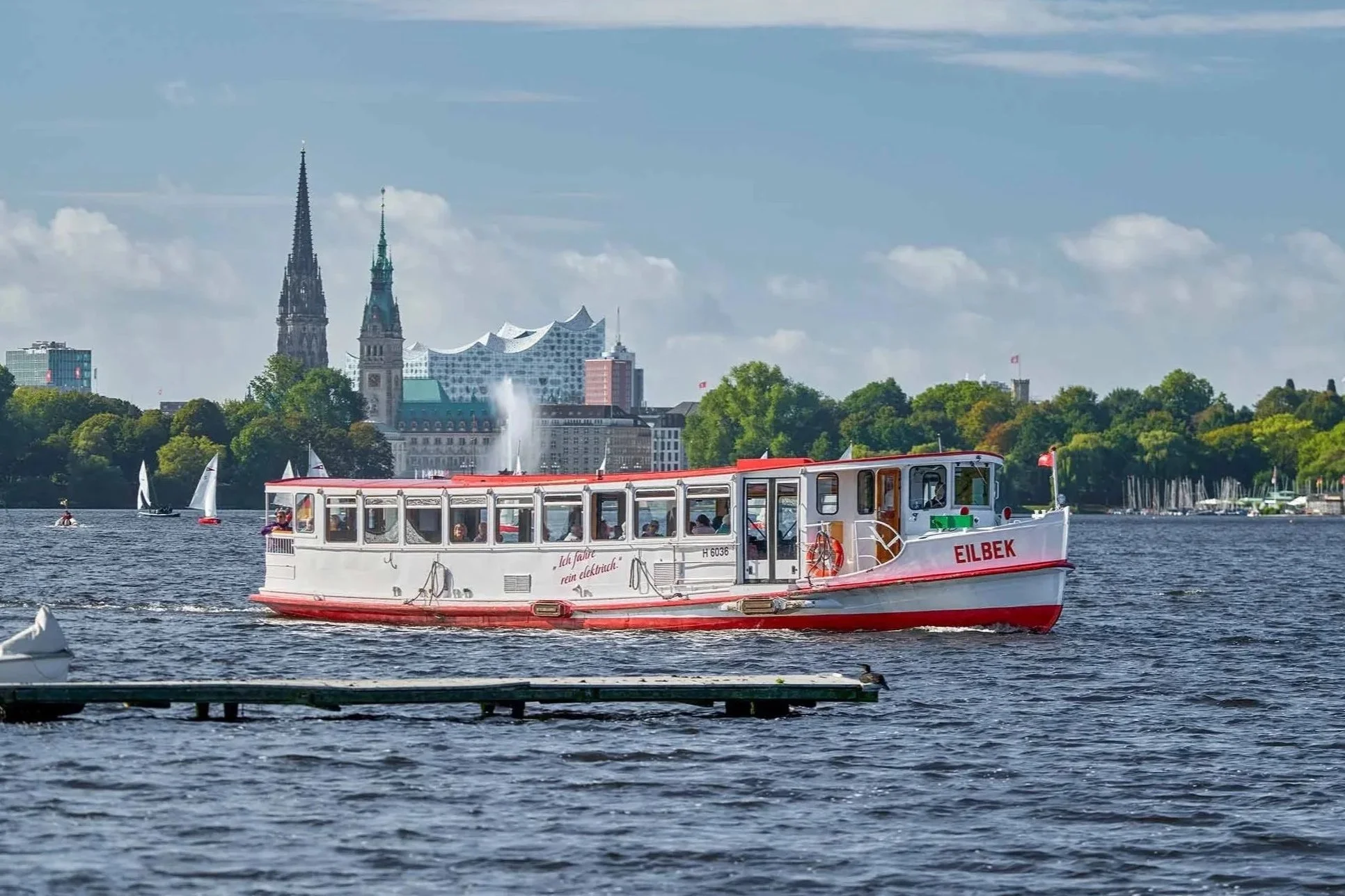 Das weiß-rote Alsterschiff Eilbek fährt über die Hamburger Außenalster. Dahinter öffnet sich der Blick auf die Hamburger Innenstadt mit markanten Kirchtürmen, Alsterfontäne und der Elbphilharmonie am Horizont. Segelboote und die bewegte Wasseroberflä