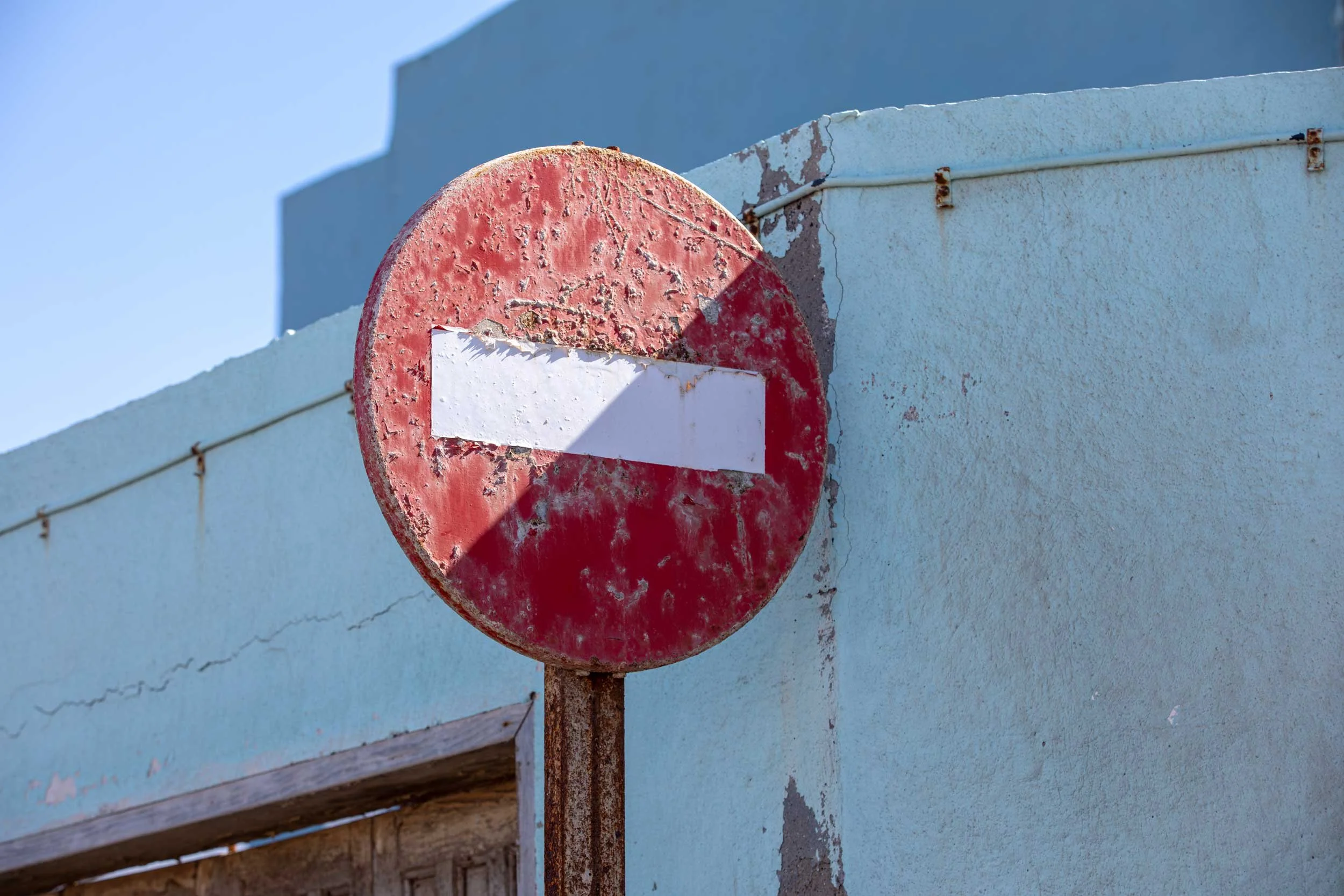 Verwittertes rundes Verkehrsschild mit weißem Querbalken an einer hellblauen Hauswand auf Gran Canaria