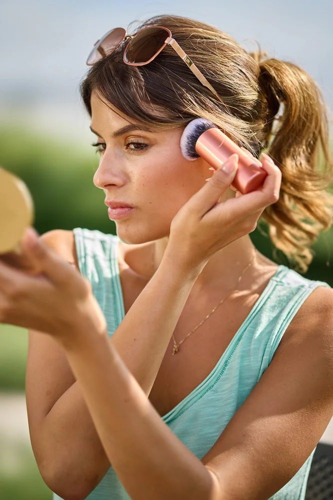 Frau trägt mit einem Make-up-Pinsel Produkt auf die Wange auf und blickt dabei in einen Handspiegel im Freien.