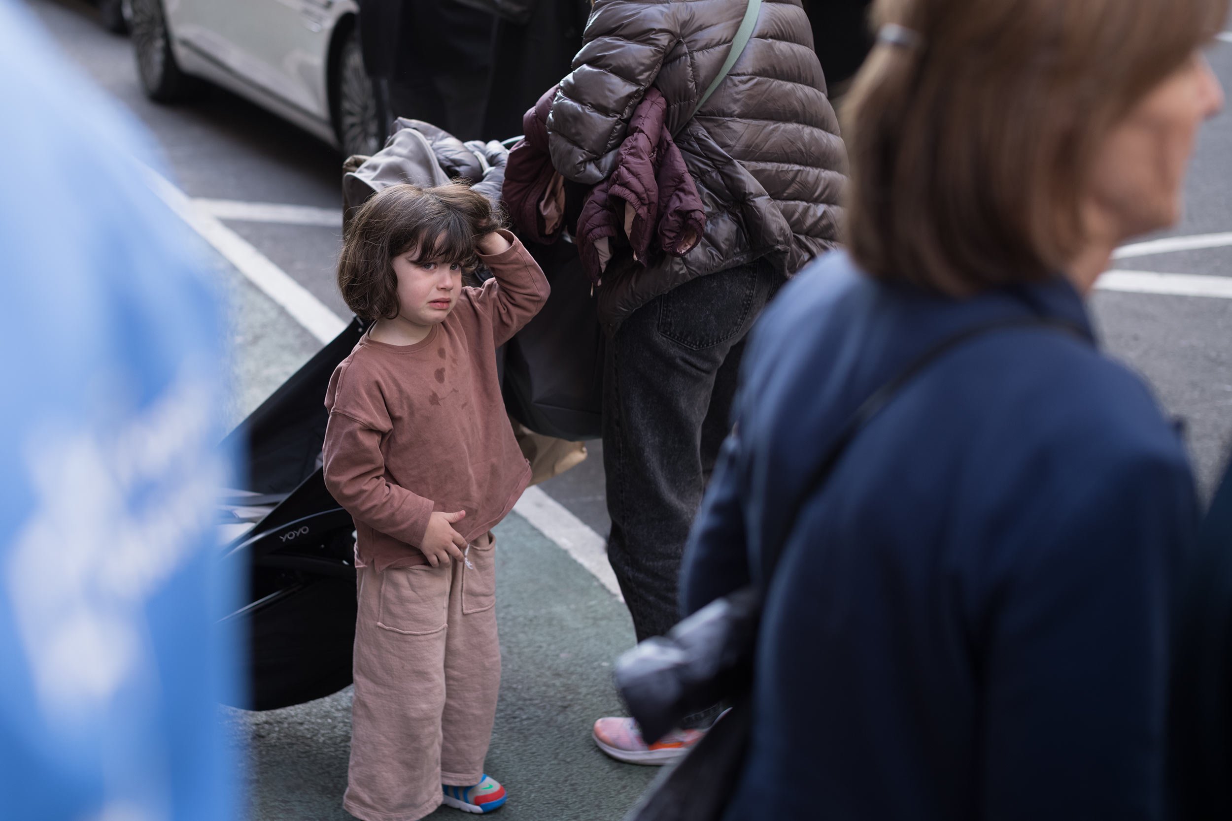 Ein kleines Mädchen in rosabrauner Kleidung steht zwischen vorbeigehenden Menschen am Straßenrand und hält eine Hand an den Kopf.